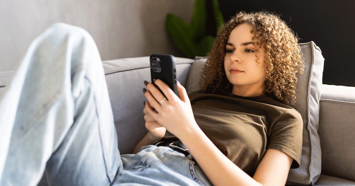 A woman relaxing on a couch while looking at her phone and researching when to get an ultrasound in early pregnancy before making decisions about her next steps.